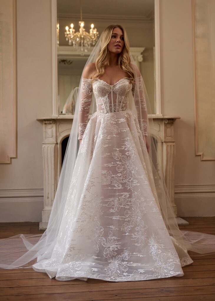 Step into a fairytale with the “Enchanted Bloom” gown. This romantic A-line ball gown features an off-the-shoulder sheer corset bodice and voluminous skirt, all adorned with intricate floral lace appliqué. A flowing cathedral-length veil completes the dreamy look, perfect for a grand entrance.